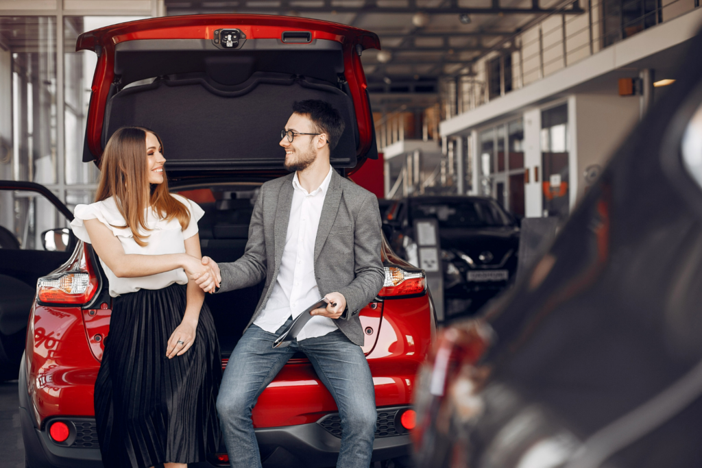 Pareja de clientes cerrando un acuerdo de compraventa con un asesor, sentados en la parte trasera de un coche rojo con el maletero abierto.