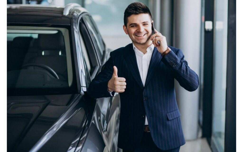 Ejecutivo de ventas con traje sonríe y da el visto bueno mientras habla por teléfono junto a un vehículo de la flota